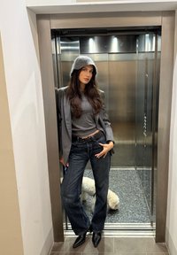 Graue Wollmütze, graues Oberteil, gestreifter Blazer, dunkle Jeans mit weitem Bein, schwarze spitze Absätze; Aufzugshintergrund mit grauen und metallischen Akzenten.