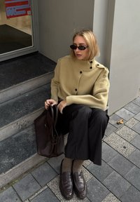 Pletený béžový oversized svetr s vysokým límcem a knoflíky, černé kalhoty s tenkými pruhy, hnědé kožené mokasíny, hnědá kabelka, sedící na kamenných schodech.