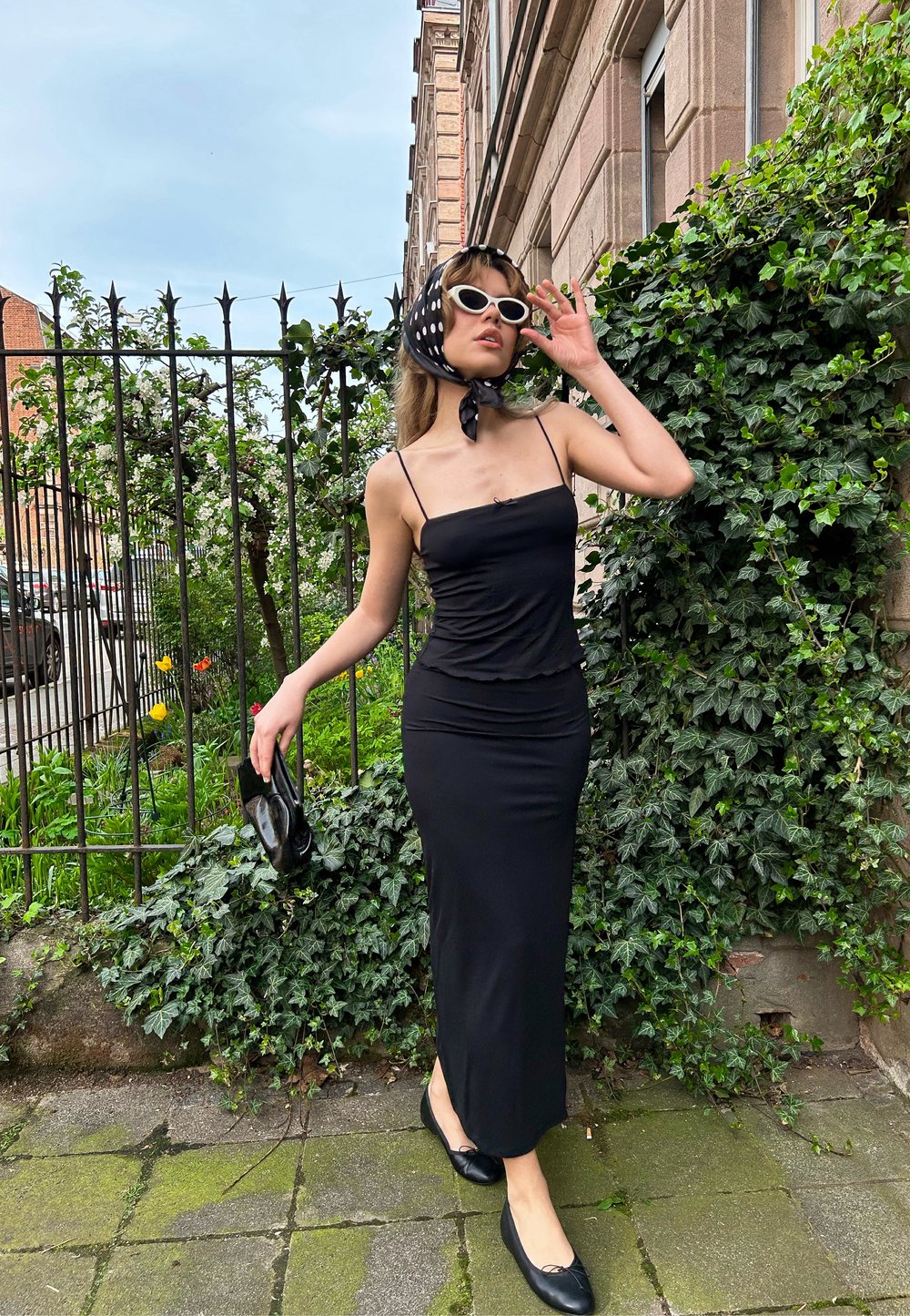 Μαύρο φόρεμα με τιράντες και εφαρμοστή γραμμή, υφασμάτινη υφή και φουντωτό στρίφωμα. Μαύρες μπαλαρίνες και μαντήλι με μαύρες και λευκές βούλες.