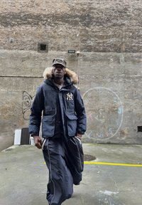 Námornická parka s kožešinovým límcem, velké khaki kapsy, vyšívané logo, v kombinaci s volnými černými kalhotami s bílými postranními pruhy. Městské pozadí.