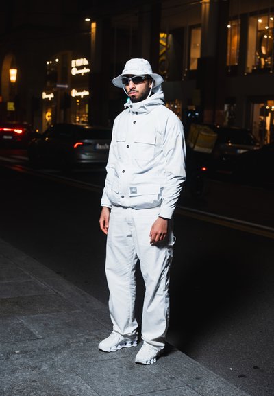 Conjunto exterior blanco que incluye una chaqueta con múltiples bolsillos y capucha, combinado con pantalones y zapatos a juego, en un fondo urbano oscuro.