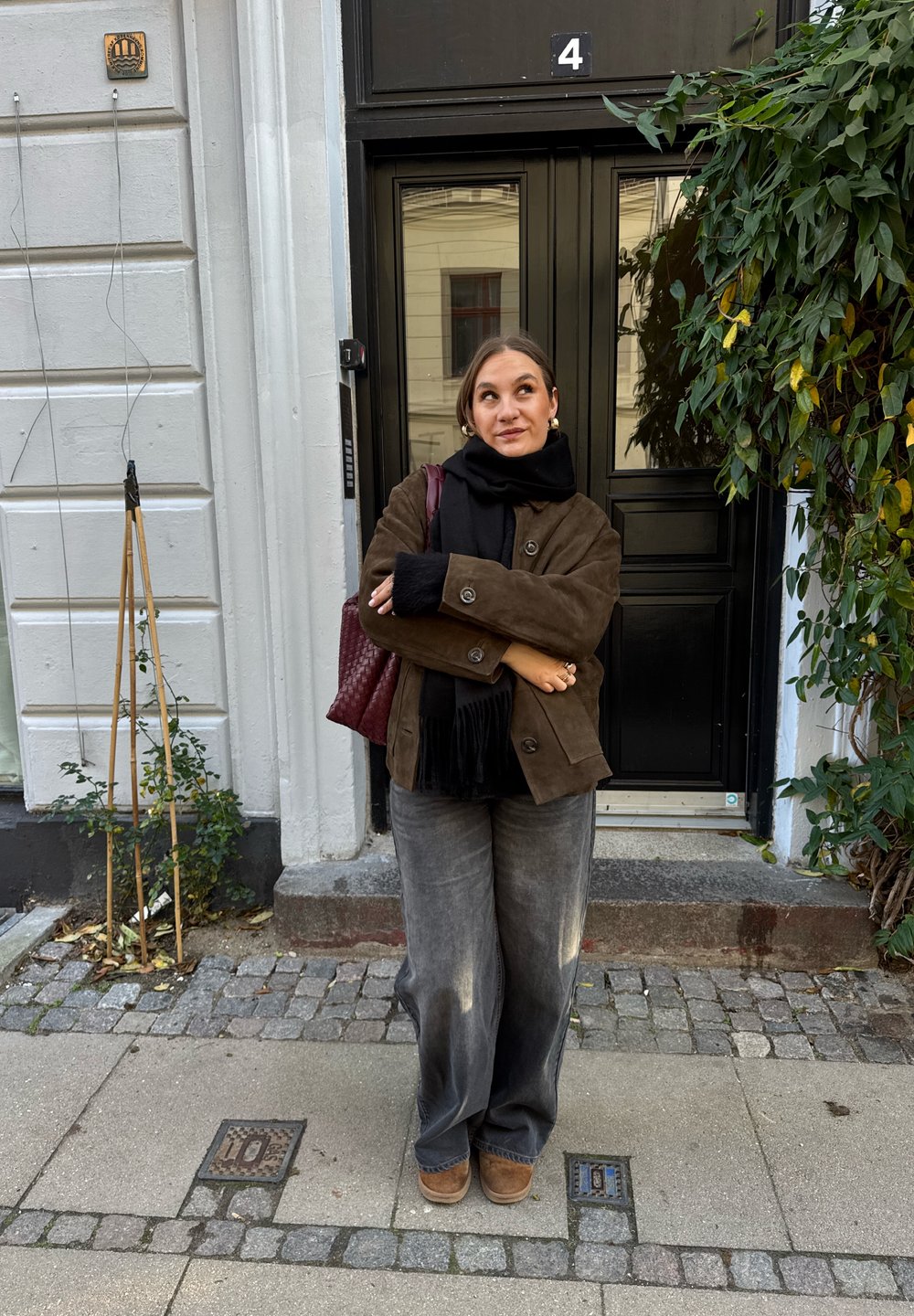 Brown suede jacket, black scarf, grey wide-leg trousers, and brown boots. Holds a burgundy handbag. Background features a black door and greenery.