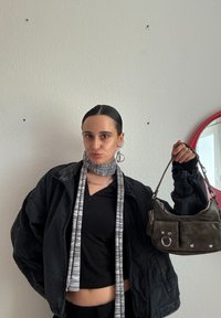 Crna skraćena jakna, sivi šal s prugastim uzorkom, mala tamna torbica s srebrnim detaljima i džepovima, jednostavna pozadina.