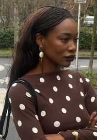 Haut marron à manches longues avec des pois blancs, associé à des boucles d'oreilles audacieuses dorées et blanches. Sac à bandoulière noir avec des bretelles visibles. Cheveux naturels.