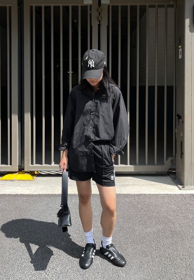Camisa de cuadros negra de botones, pantalones cortos negros con logo blanco, gorra negra, calcetines blancos y zapatillas deportivas negras con tres franjas.