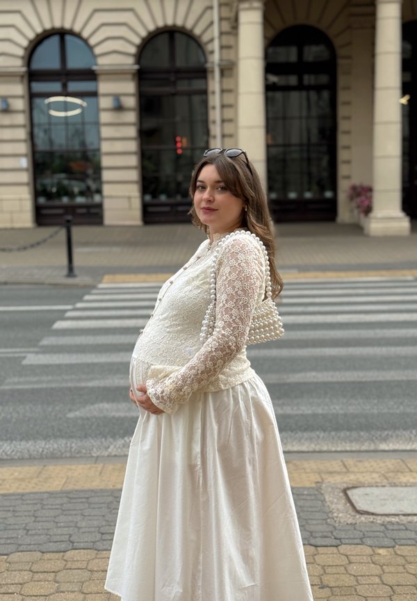 Kobieta w ciąży w białej koronkowej sukience i z torebką perłową stoi przy przejściu dla pieszych z historycznym budynkiem w tle.