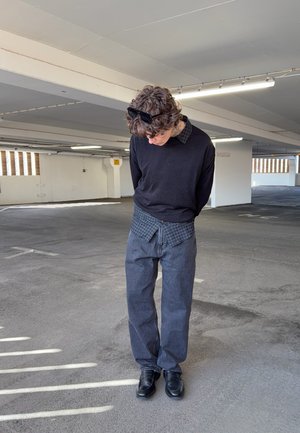 Joven con cabello rizado y gafas de sol sobre la cabeza, de pie mirando hacia abajo en un estacionamiento mayormente vacío, vistiendo ropa casual en capas.