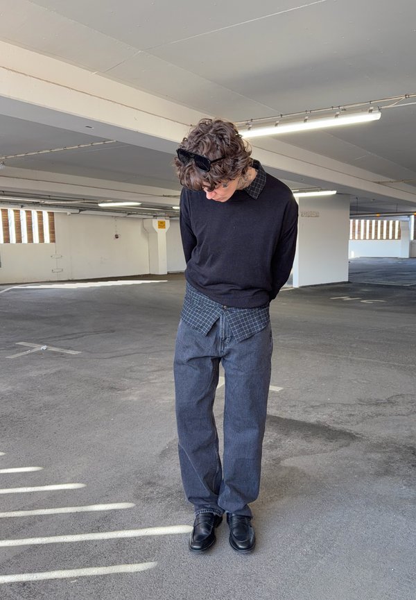 Giovane con capelli ricci e occhiali da sole sulla testa che guarda verso il basso in un parcheggio quasi vuoto, indossando abbigliamento casual a strati.