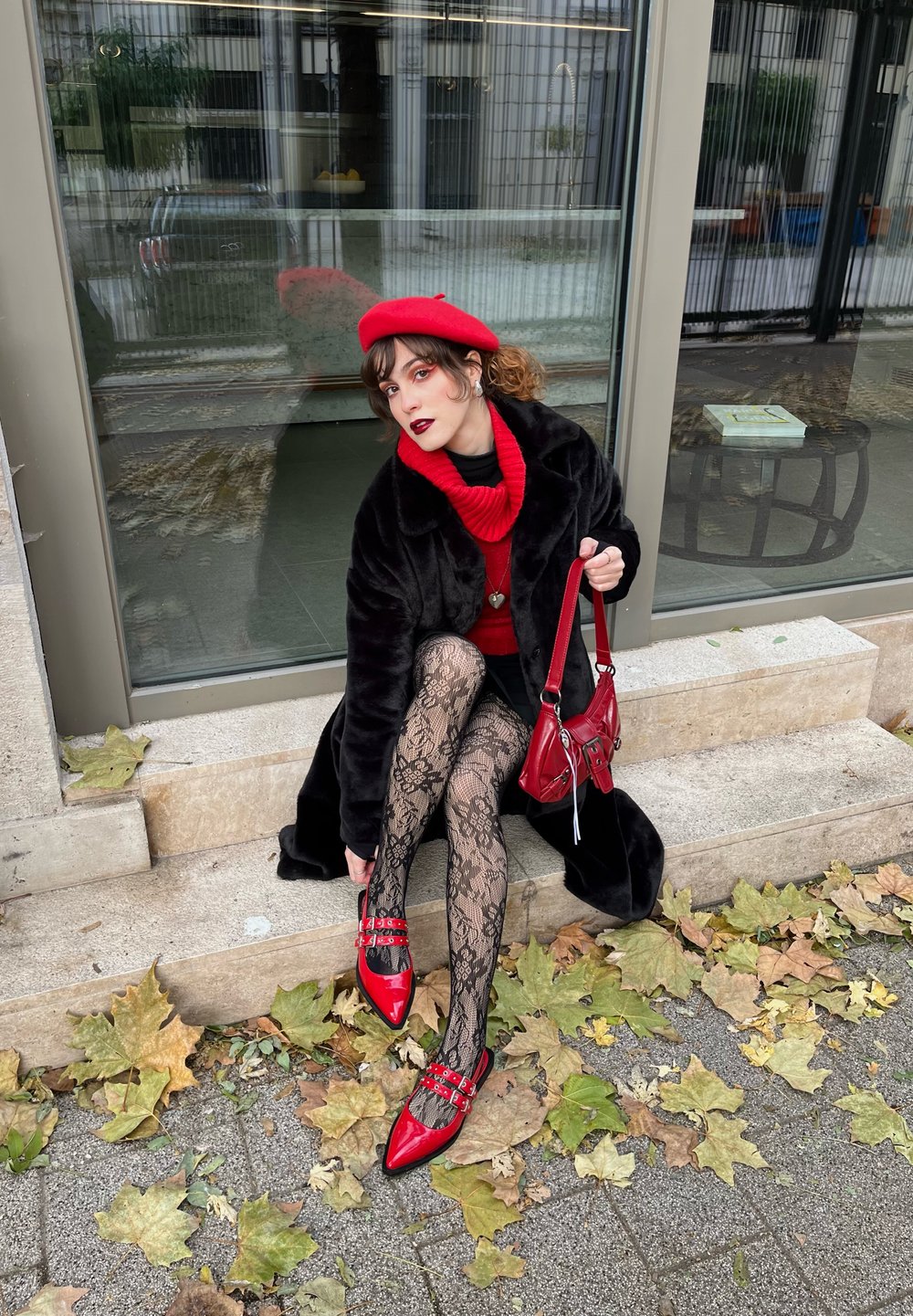 Rosso tacchi alti con cinturini, calze di pizzo nere, maglione rosso e cappotto nero. Seduta su gradini di pietra circondata da foglie autunnali.