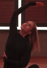 Femeie îmbrăcată într-un top negru cu mânecă lungă, efectuând o întindere laterală cu un braț ridicat deasupra capului și privind în sus, într-o cameră cu panouri din lemn.