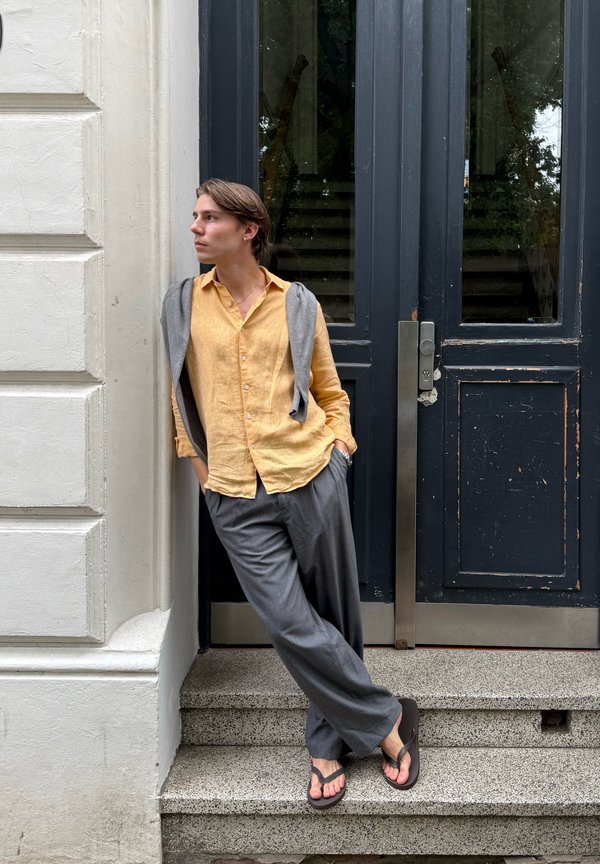 Ung person med kort hår, der læner sig op ad en hvid væg, iført en gul skjorte, grå bukser, grå sweater over skuldrene og sorte flip-flops.
