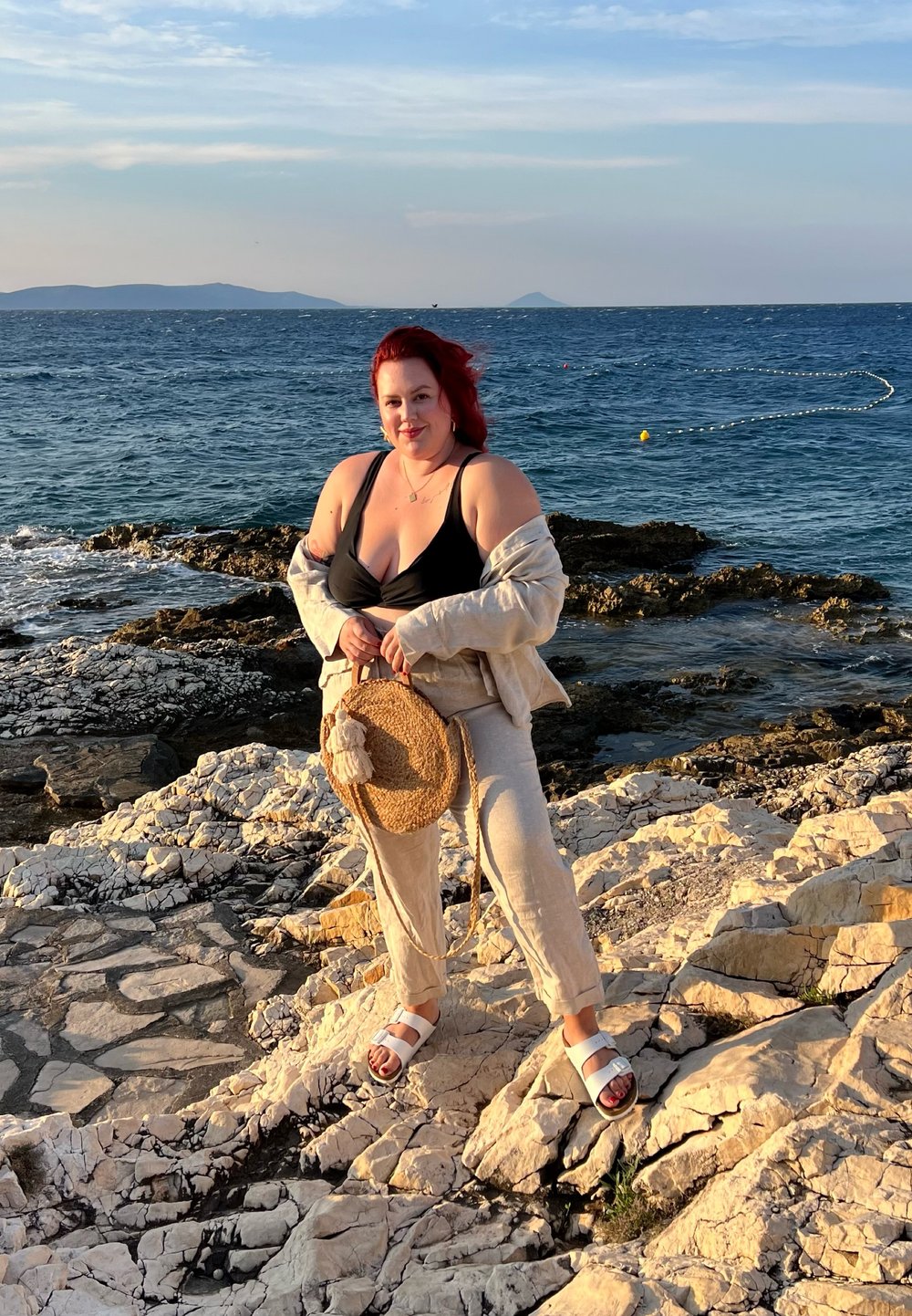 Kobieta stoi na skalistym wybrzeżu, ubrana w czarną bluzkę i jasne lniane spodnie, trzymając okrągłą słomianą torbę i nosząc sandały. W tle widać ocean.
