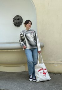 Ριγέ μακρυμάνικη μπλούζα, ασπρόμαυρη, με κόκκινο λογότυπο. Μπλε τζιν, μαύρα αθλητικά π�απούτσια, και μια λευκή τσάντα tote με κόκκινο κείμενο και σχέδιο.