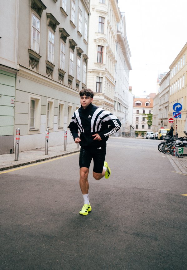 Νέος άνδρας με ασπρόμαυρη αθλητική ένδυση και παπούτσια νέον τρέχει σε άδεια αστική οδό περιτριγυρισμένη από ιστορικά κτίρια.