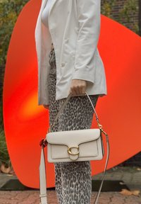 Světlé béžové kabelky se zlatým uzávěrem, texturovanou kůží a odnímatelným ramenním popruhem, držené proti jasně oranžovému pozadí.