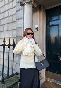 Femme en gants blancs, écharpe et veste, souriant auprès d'une colonne en pierre et d'une porte noire, tenant un sac à main noir dans une rue de la ville.