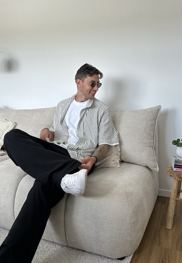 Homme portant des lunettes de soleil, des baskets blanches et une chemise rayée, assis en tailleur sur un canapé beige dans un salon minimaliste.