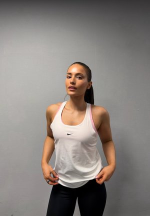 Mujer joven con cabello castaño recogido en una coleta, lleva una camiseta sin mangas blanca de Nike y leggings negros, de pie frente a una pared gris lisa.