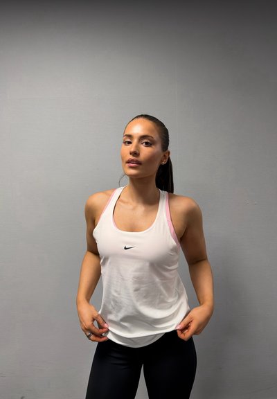 Mujer joven con cabello castaño recogido en una coleta, lleva una camiseta sin mangas blanca de Nike y leggings negros, de pie frente a una pared gris lisa.