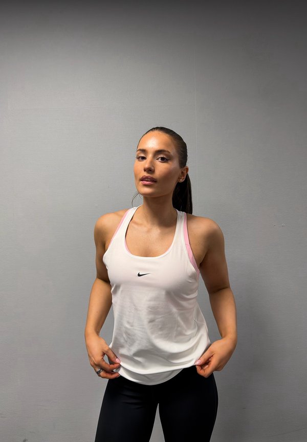 Jeune femme aux cheveux bruns attachés en queue de cheval, portant un débardeur blanc Nike et un legging noir, debout devant un mur gris uni.
