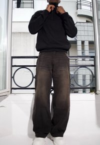 Sweatshirt noir zippé avec col haut, associé à un pantalon ample marron foncé. Des baskets blanches complètent le look décontracté.