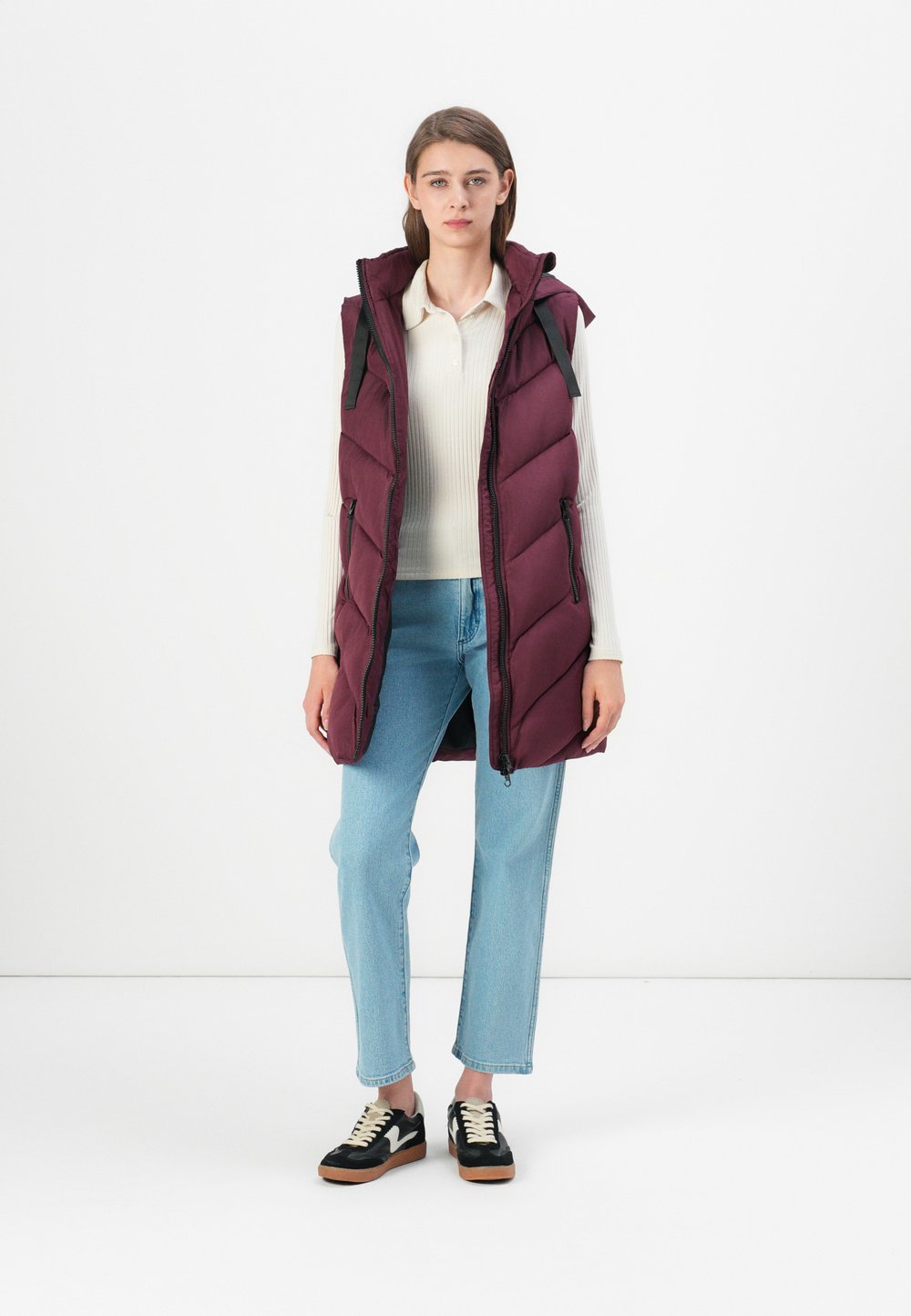 Σκούρο μοβ γιλέκο τύπου puffer με κουκούλα, φερμουάρ μπροστά και οριζόντιες ραφές. Φοριέται πάνω από ένα ανοιχτόχρωμο τοπ και συνδυάζεται με ανοιχτό μπλε τζιν.