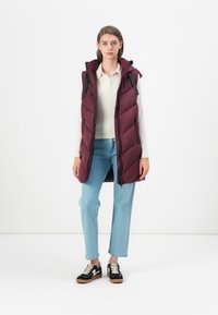 Σκούρο μοβ γιλέκο τύπου puffer με κουκούλα, φερμουάρ μπροστά και οριζόντιες ραφές. Φοριέται πάνω από ένα ανοιχτόχρωμο τοπ και συνδυάζεται με ανοιχτό μπλε τζιν.