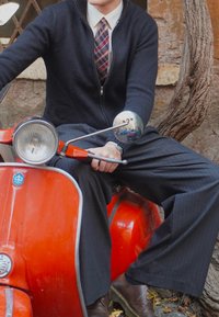 Sarkans skūteris ar hromētiem akcentiem, caurspīdīga luktura, un apaļa spogulis. Vīrietis tumši zilā džemperī un svītrainās biksēs sēž pārliecinoši, rokas uz stūres.