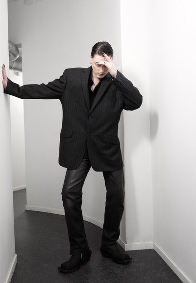 Persona con una blazer negra de gran tamaño y pantalones de cuero apoyada contra una pared, con la cabeza apoyada en la mano, de pie en un pasillo estrecho.