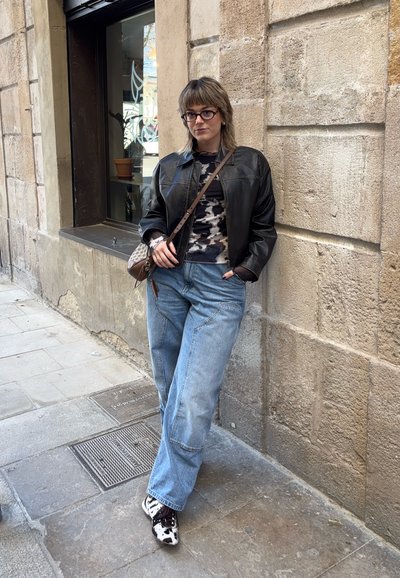 Chaqueta de cuero negra sobre una blusa de camuflaje, vaqueros azules holgados y zapatillas con patrón en blanco y negro. Bandolera marrón, de pie junto a una pared.