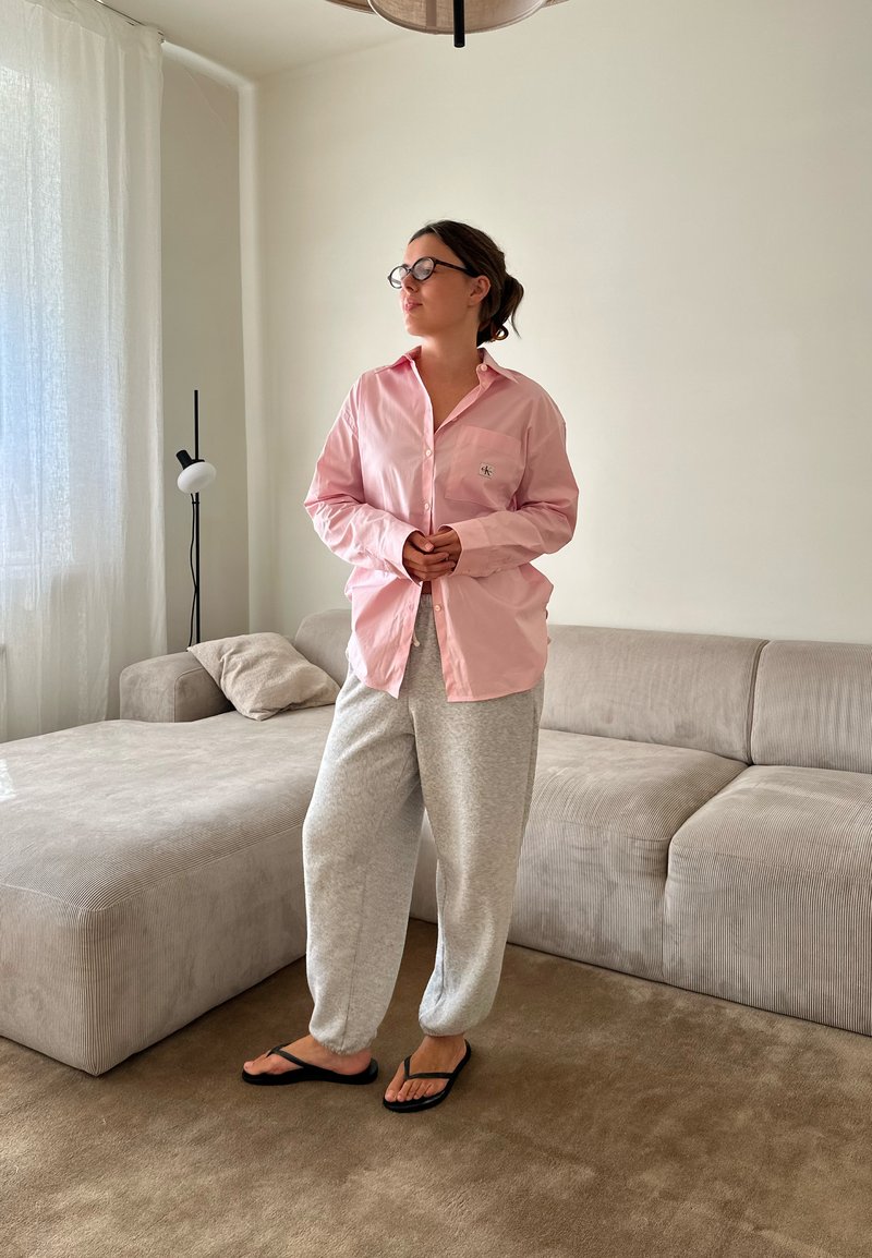 Camisa ligeiramente rosa com botão e bolso no peito, calças de treino cinzentas e chinelos pretos. Texturas suaves num ambiente interior minimalista.