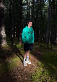 Hanorac verde cu un design grafic, pantaloni scurți negri, șosete albe și adidași gri, stând pe un sol de pădure acoperit cu ace de pin.