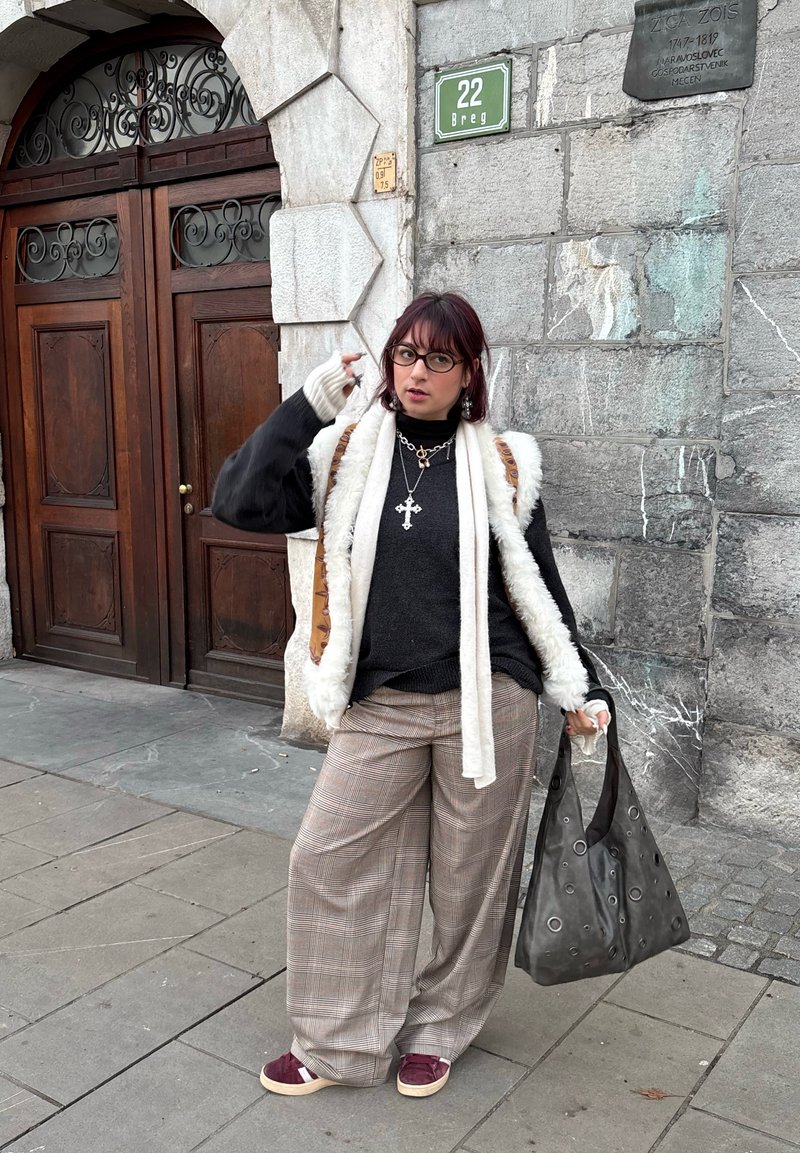 Pull en laine gris superposé avec un gilet en fourrure blanc, pantalon large à carreaux, baskets bordeaux et sac gris à motif. Debout contre un mur en pierre.