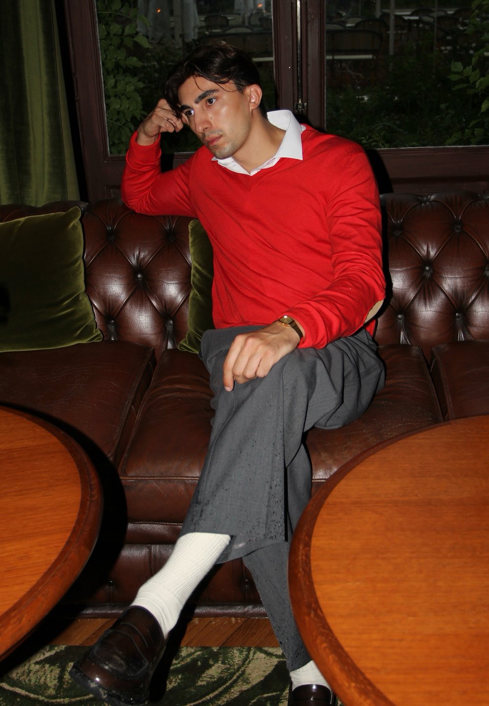 Roter Pullover mit weißem Kragen, graue Hose, schwarze Slipper, sitzend auf einem dunkelbraunen Ledersofa, mit Holztischen und grünen Akzenten.