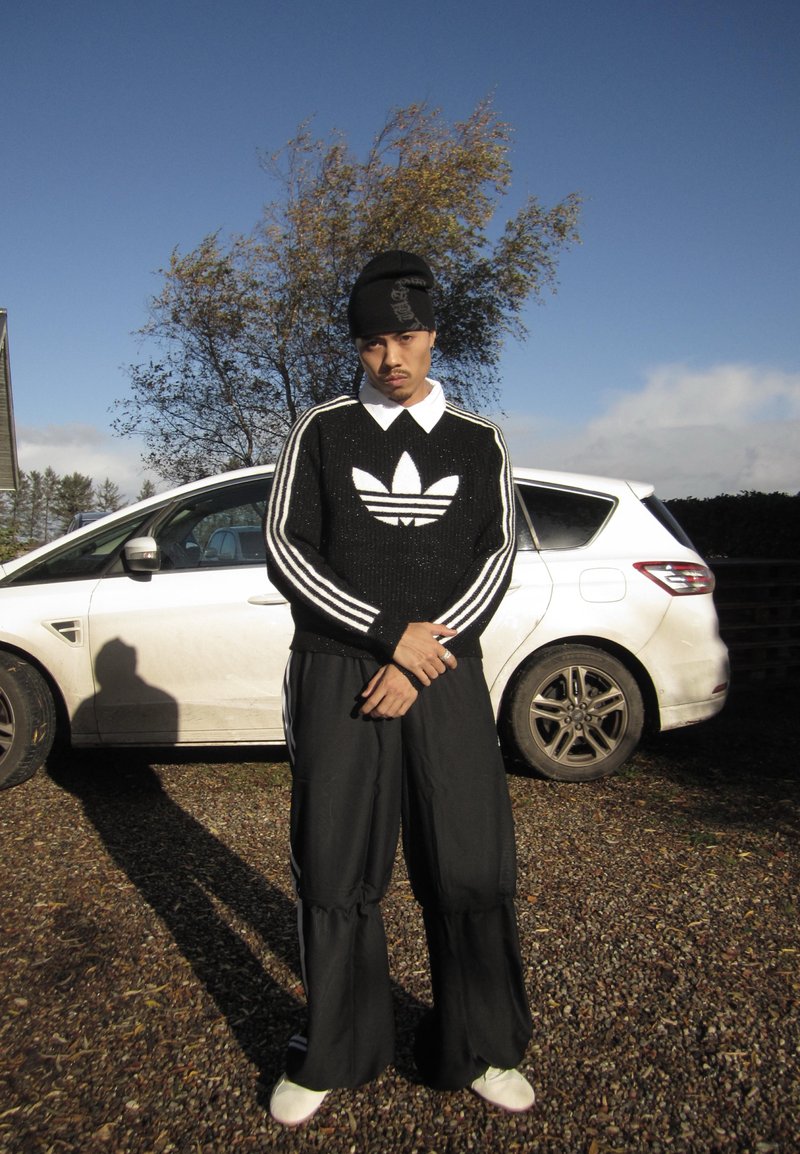 Uomo con felpa nera Adidas, pantaloni larghi neri e scarpe bianche, in piedi con le braccia incrociate davanti a un'auto bianca su ghiaia sotto un cielo limpido.