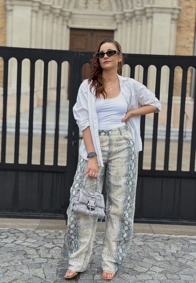 Mujer con camiseta blanca sin mangas, camisa blanca suelta, pantalones de serpiente, gafas de sol, sosteniendo un bolso gris, de pie sobre adoquines cerca de una puerta negra.