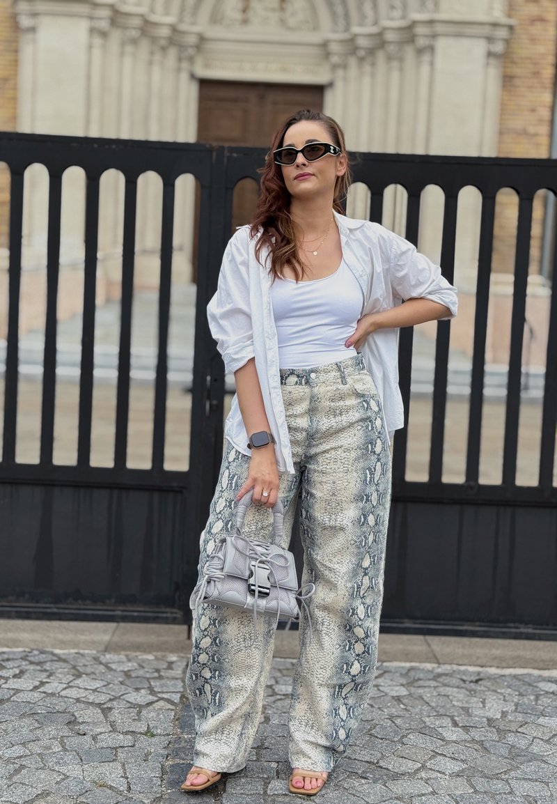 Femme portant un débardeur blanc, une chemise blanche ample, un pantalon imprimé serpent, des lunettes de soleil, tenant un sac à main gris, se tenant sur des pavés près d'un portail noir.