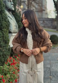 Hnědý pletený kardigan, krémová pletená šála, světle béžové oblečení, stříbrná kabelka s řetízkovými popruhy, venkovní prostředí se keři a květinami.