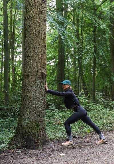 Una persona se estira contra un tronco de árbol texturizado en un bosque verde y frondoso, vistiendo una chaqueta negra, leggings y zapatos con suela de color claro.