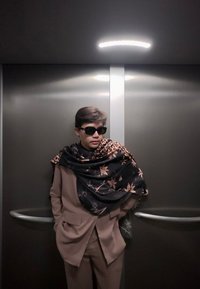 Personne portant des lunettes de soleil, une écharpe à motifs floraux et léopard, et un costume marron, debout les mains dans les poches à l'intérieur d'un ascenseur métallique.