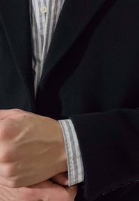 Άνδρας φοράει μαύρο παλτό πάνω από ριγέ πουκάμισο με κουμπιά, προσαρμόζοντας την μανσέτα με το ένα χέρι να κρατάει τον αντίθετο καρπό.