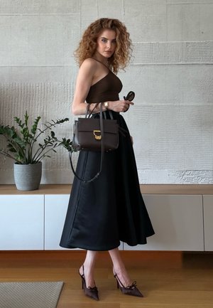 Top marrón sin mangas, falda midi negra, bolso oscuro con herrajes dorados y tacones de punta marrones. Cabello rizado y gafas de sol en la mano.