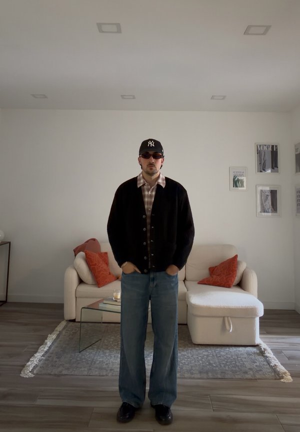 Moški, oblečen v črni pulover z gumbi, karo srajco, modre kavbojke, sončna očala in črno kapo, stoji v minimalistični dnevni sobi z bež kavčem in oranžnimi blazinami.
