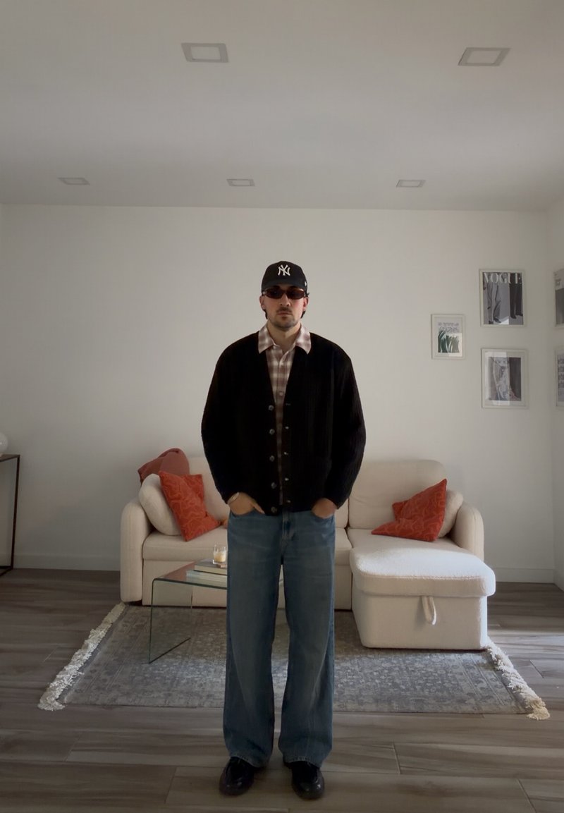 Mand iført sort cardigan, ternet skjorte, blå jeans, solbriller og sort kasket, stående i et minimalistisk stue med beige sofa og orange puder.