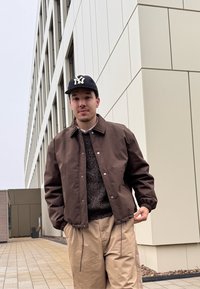 Veste coupe-vent marron avec poignets élastiques et boutons-pression, portée sur un pull sombre, associée à un pantalon beige. Fond de bâtiment neutre.