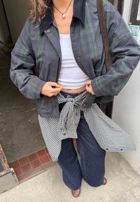 Karo, temno zelena in črna jakna, bela majica brez rokavov, karirasta srajca zavezana okoli pasu, širše kavbojke, rjave čevlje, stoji na pločniku.