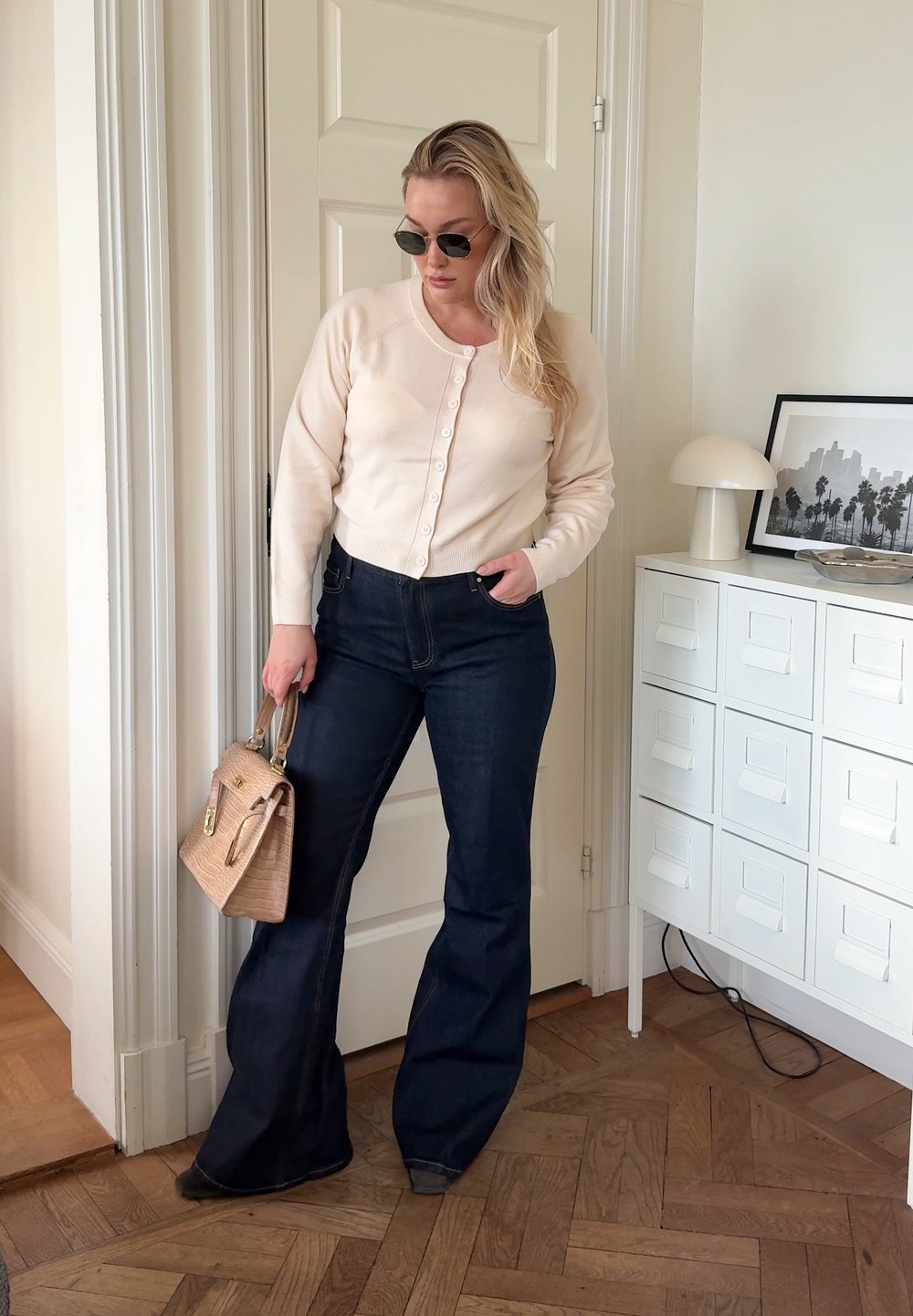 Beige kofta med knappar, mörka högmidjade utsvängda jeans och en strukturerad tanfärgad handväska. Runda solglasögon fulländar outfiten.