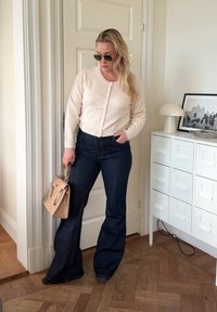 Beige kofta med knappar, mörka högmidjade utsvängda jeans och en strukturerad tanfärgad handväska. Runda solglasögon fulländar outfiten.