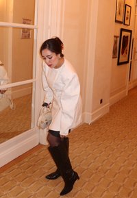 Giacca bianca oversize con dettagli ai bottoni, abbinata a una mini gonna nera e stivali in pelle neri fino al ginocchio. Tiene una piccola borsa.