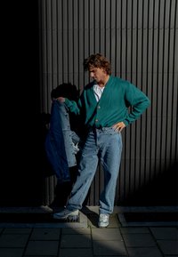 Türkiser Strickcardigan mit Holzknöpfen, über einem weißen Hemd getragen. Helle blaue Jeans und eine Jeansjacke in der Hand, vor einem dunklen Hintergrund.